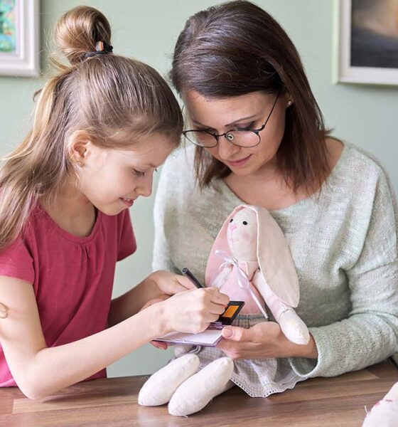 Mother and daughter child play together at home, girl and mom make make-up paints cheeks to toy handmade doll bunny sewn on sewing machine